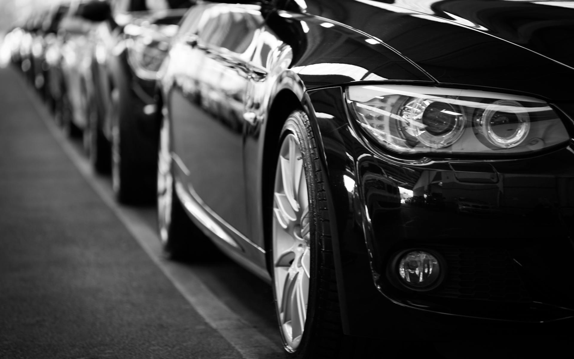 Imagem de uma fila de carros sedans pretos com reflexos da
          iluminação natural, todos desligados e alinhados a uma faixa branca
          no chão.
